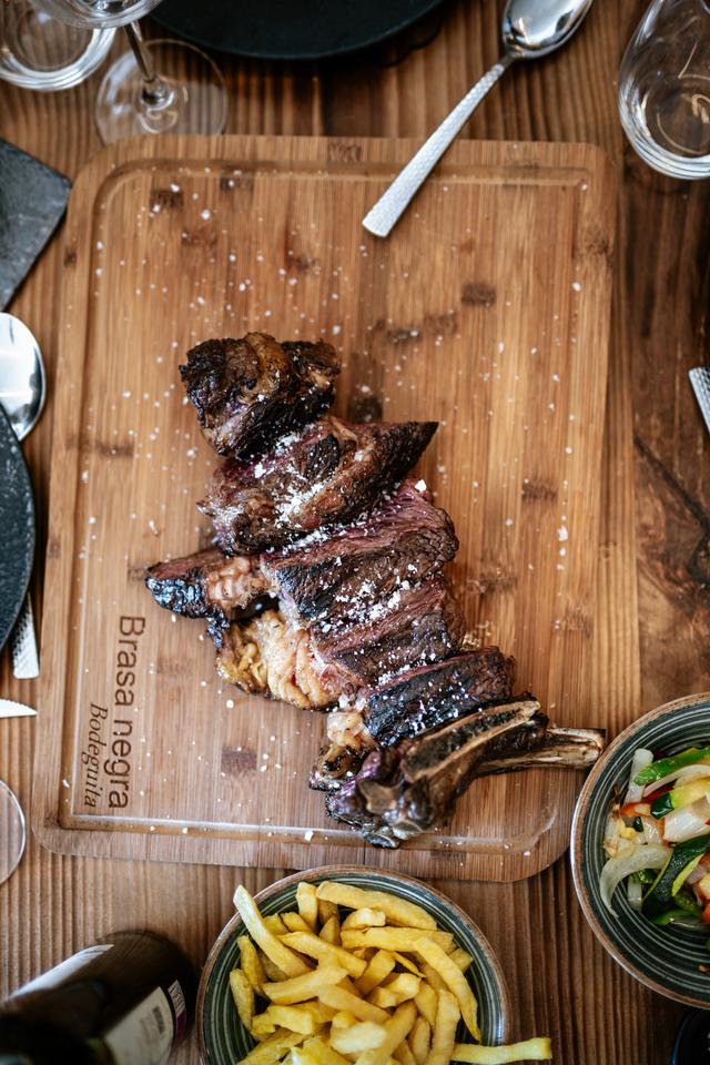 Carne a la brasa cortada sobre una tabla grabada con el nombre Brasa Negra, con patatas y ensalada